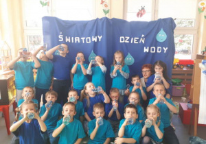 Grupa dzieci pozuje do zdjęcia pijąc wodę z butelek. W tle dekoracja z napisem "Światowy Dzień Wody".